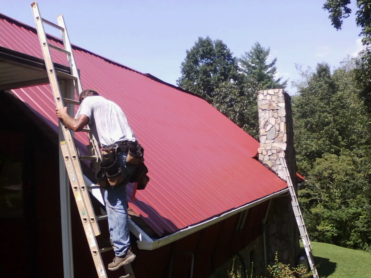 Roofer on a ladder beside a red metal roof and stone chimney — Soffit & Fascia Repair by Roofing Brothers in Stanleytown